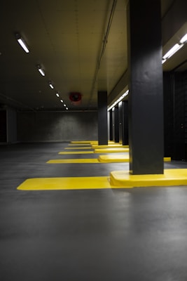 A dimly lit parking garage featuring multiple parking spaces marked by bright yellow lines. The concrete ceiling and floor have a gray hue, complemented by black support columns. Overhead fluorescent lights provide limited illumination, casting a subdued atmosphere.