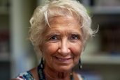 An older woman with short, curly white hair, wearing earrings and a colorful patterned top, smiles warmly. The background is softly blurred, emphasizing her facial features and expression.