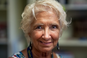 An older woman with short, curly white hair, wearing earrings and a colorful patterned top, smiles warmly. The background is softly blurred, emphasizing her facial features and expression.