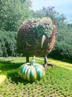 A garden sculpture made from plants shaped like a bird with a prominent beak stands on a patch of vibrant green grass. In front of it, a large, decorative pumpkin with swirling colors of blue, orange, and green adds a whimsical touch to the scene. Dense bushes and trees form a lush, green backdrop.