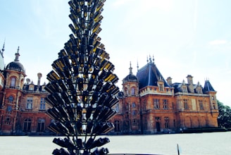 A large, ornate building with intricate architectural details stands in the background. In the foreground, a unique sculpture made of stacked green glass bottles rises vertically, resembling a tree. The sky is clear and bright, casting a warm light over the scene.