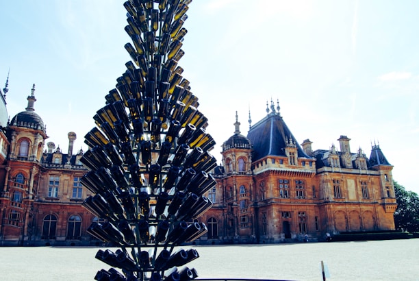 A large, ornate building with intricate architectural details stands in the background. In the foreground, a unique sculpture made of stacked green glass bottles rises vertically, resembling a tree. The sky is clear and bright, casting a warm light over the scene.