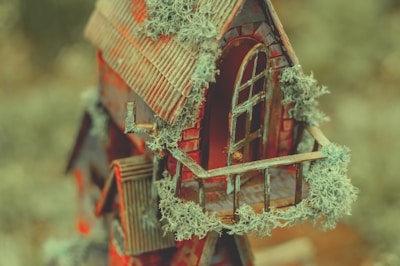 A miniature house with a rustic, weathered appearance, featuring a sloped roof and detailed wooden balcony. The house is adorned with patchy green mosses or lichens, adding to its aged look. The materials appear to be crafted from rough, textured wood and paint, with an arched doorway and window.