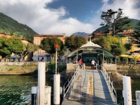 A quaint lakeside town with colorful buildings flanked by lush green trees. A dock extends over the water, leading to a small building with the sign 'Bellano'. People are casually walking along the dock, and the backdrop consists of green hills and partly cloudy skies.