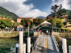 A quaint lakeside town with colorful buildings flanked by lush green trees. A dock extends over the water, leading to a small building with the sign 'Bellano'. People are casually walking along the dock, and the backdrop consists of green hills and partly cloudy skies.