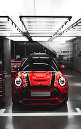 A bright red Mini John Cooper Works car is showcased in a modern, well-lit garage or showroom. The car's sleek design features prominent black racing stripes running over the hood.