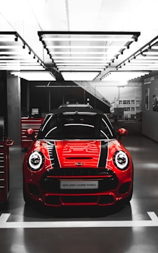 A bright red Mini John Cooper Works car is showcased in a modern, well-lit garage or showroom. The car's sleek design features prominent black racing stripes running over the hood.