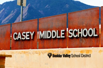 A large rust-colored metal sign displays the words 'Casey Middle School' with bold white lettering. Below, another sign indicates it is part of the Boulder Valley School District. In the background, large mountain peaks are visible, suggesting a natural setting.