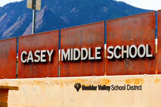 A large rust-colored metal sign displays the words 'Casey Middle School' with bold white lettering. Below, another sign indicates it is part of the Boulder Valley School District. In the background, large mountain peaks are visible, suggesting a natural setting.