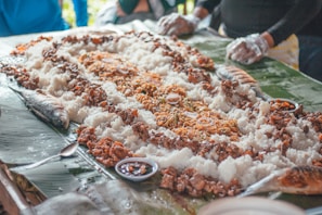 A traditional communal meal is spread out on banana leaves featuring white rice surrounded by various dishes, including grilled fish, pieces of meat, noodles, and small bowls of sauce. People with gloves are seen around the table, engaging in this shared eating experience.