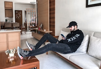 A person is lounging on a beige sofa in a modern living room wearing a black hoodie and cap, looking at a smartphone. The room features light wooden furniture, a wooden partition, and a table with small decorative items and a plant. The background includes a dining area and a door adorned with stickers.