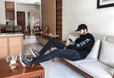 A person is lounging on a beige sofa in a modern living room wearing a black hoodie and cap, looking at a smartphone. The room features light wooden furniture, a wooden partition, and a table with small decorative items and a plant. The background includes a dining area and a door adorned with stickers.