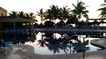A serene swimming pool area with blue sun loungers arranged around it. Palm trees line the background, silhouetted against a vibrant sunset sky. The pool reflects the colors of the sunset, creating a calming ambiance. A tiled roof structure is visible on the left side.