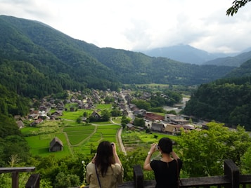 Two people stand on an observation deck capturing the view of a picturesque village nestled in a lush green valley surrounded by mountains. Traditional wooden houses with steep thatched roofs are scattered throughout the landscape, with vibrant green rice paddies in the foreground. The sky is partly cloudy, casting a serene and tranquil atmosphere over the entire scene.