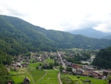 A picturesque panoramic view of a rural village with traditional wooden houses nestled in a lush, green valley surrounded by dense forested mountains. The village is characterized by its harmonious integration with the natural landscape, and a river runs alongside it, enhancing the idyllic setting.