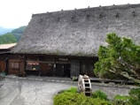 A traditional wooden building with a thatched roof is set in a rural environment. The structure has a rustic appearance and features a small wooden waterwheel in front. The surrounding landscape includes lush greenery and trees, creating a serene and natural atmosphere.