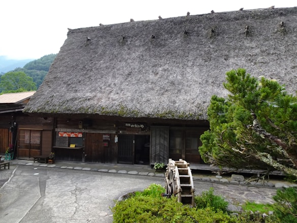 A traditional wooden building with a thatched roof is set in a rural environment. The structure has a rustic appearance and features a small wooden waterwheel in front. The surrounding landscape includes lush greenery and trees, creating a serene and natural atmosphere.