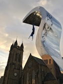 A dynamic sculpture of a diver hangs upside down from a large, curved metallic structure with engraved patterns. The background features an old stone church with intricate architectural details, including a tower and pointed spires. The sky exhibits soft hues, creating a striking contrast with the modern art piece.