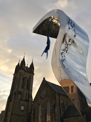 A dynamic sculpture of a diver hangs upside down from a large, curved metallic structure with engraved patterns. The background features an old stone church with intricate architectural details, including a tower and pointed spires. The sky exhibits soft hues, creating a striking contrast with the modern art piece.