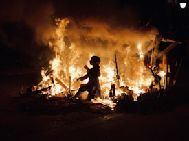 A scene featuring a large bonfire with flames reaching upward and surrounding several figurines or statues. The setting appears to be outdoors at night, with a dark background highlighting the fire's intensity and brightness.