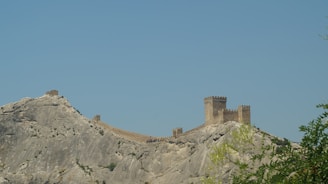 An ancient fortress with towers and connecting stone walls is perched on a rocky hillside. The structure appears weathered and historical, with the walls following the contours of the rugged terrain. The sky above is clear, and greenery can be seen in the foreground, suggesting a serene, isolated location.