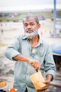 An older man with a grey beard is stirring a plastic container of food. He wears a light blue shirt and stands outdoors, with a blurred background featuring people and structures.
