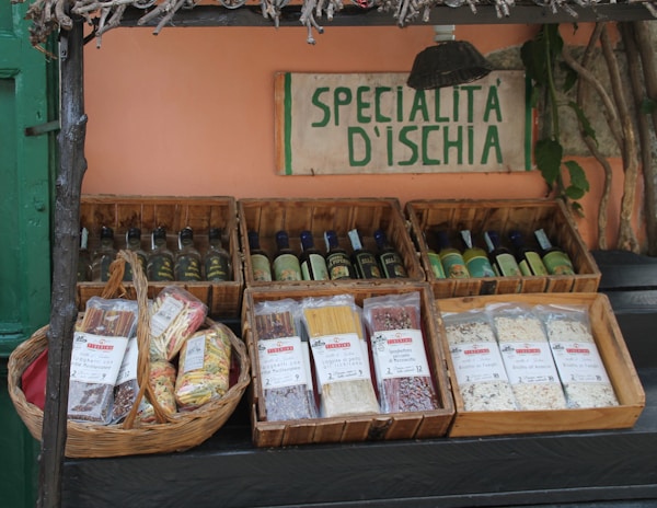 A display of various Italian specialty products, including packaged pasta and bottles of liqueur, arranged in wooden crates and a basket. A hand-painted sign reading 'Specialità D'Ischia' is placed prominently above the products. The setup suggests an artisanal or rustic market ambiance, with some greenery and wooden elements adding to the aesthetic.