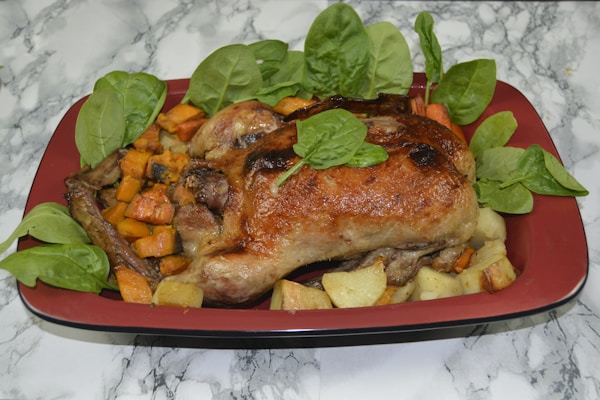 A roasted chicken is placed in a rectangular red dish surrounded by roasted potatoes, carrots, and garnished with fresh spinach leaves. The dish is set against a marble surface.
