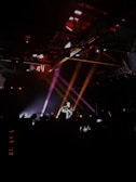 A dimly lit concert scene with a performer playing guitar on stage, illuminated by colorful spotlights. The audience is visible as silhouettes, capturing the moment with phones.