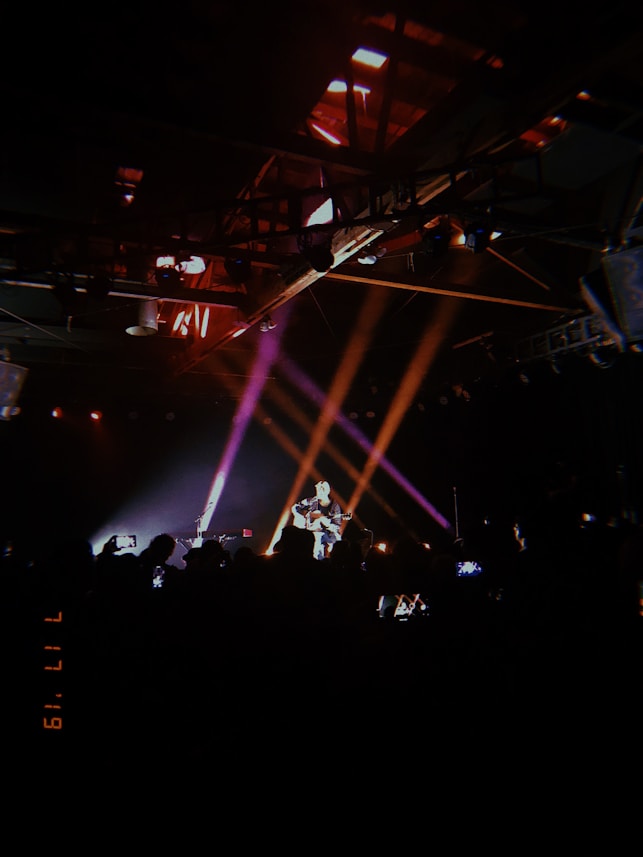 A dimly lit concert scene with a performer playing guitar on stage, illuminated by colorful spotlights. The audience is visible as silhouettes, capturing the moment with phones.