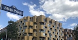 A modern residential building with a unique, staggered facade featuring numerous windows and balconies. The structure is predominantly yellow with brown accents. A street sign with the text 'Aleja Pracy' is visible in the foreground. The sky is partly cloudy, and there are green trees around.