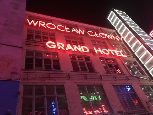 A nighttime urban scene featuring an old building with large red neon signs reading 'WROCLAW GLOWNY' and 'GRAND HOTEL' across the windows. The building facade shows some wear, and there is a modern, well-lit vertical sign structure on the right side.