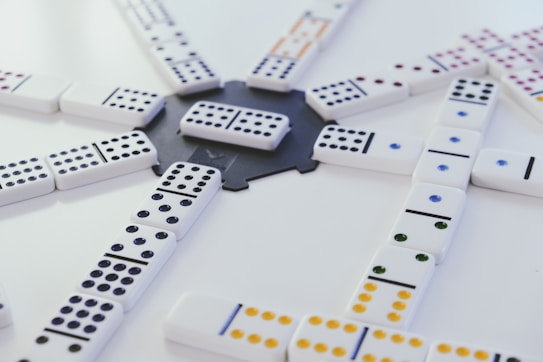 A collection of white dominoes with colored dots is arranged on a flat surface, converging at a central point. The arrangement suggests an ongoing game with multiple lines branching out, displaying a variety of dot patterns in colors like black, blue, green, and yellow.