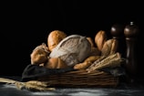 A variety of artisanal breads are placed in a wicker basket lined with a dark cloth, accompanied by ears of wheat. The scene is set against a dark, moody background with faint dustings of flour on the surface. Two wooden pepper mills stand in the background, adding a rustic touch.