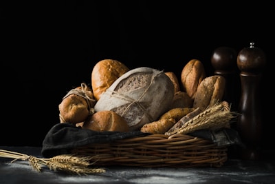 A variety of artisanal breads are placed in a wicker basket lined with a dark cloth, accompanied by ears of wheat. The scene is set against a dark, moody background with faint dustings of flour on the surface. Two wooden pepper mills stand in the background, adding a rustic touch.