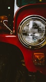 A close-up view of a vintage vehicle's headlight, featuring a large round chrome headlamp with detailed glass patterns. The vibrant red paint of the vehicle's body contrasts with the shiny metal of the headlight and the small amber-colored indicator light below.