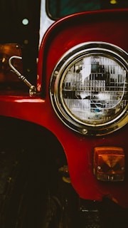 A close-up view of a vintage vehicle's headlight, featuring a large round chrome headlamp with detailed glass patterns. The vibrant red paint of the vehicle's body contrasts with the shiny metal of the headlight and the small amber-colored indicator light below.