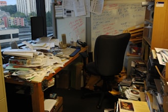 A cluttered office with a desk covered in papers, CDs, and miscellaneous items. A black office chair is positioned in front of the desk. There is a whiteboard on the wall with various notes scribbled in different colors. Shelves and boxes filled with more papers and folders surround the desk. The room has a window showcasing a cityscape with buildings in the background.