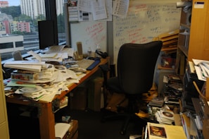 A cluttered office with a desk covered in papers, CDs, and miscellaneous items. A black office chair is positioned in front of the desk. There is a whiteboard on the wall with various notes scribbled in different colors. Shelves and boxes filled with more papers and folders surround the desk. The room has a window showcasing a cityscape with buildings in the background.