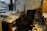 A cluttered office with a desk covered in papers, CDs, and miscellaneous items. A black office chair is positioned in front of the desk. There is a whiteboard on the wall with various notes scribbled in different colors. Shelves and boxes filled with more papers and folders surround the desk. The room has a window showcasing a cityscape with buildings in the background.
