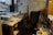 A cluttered office with a desk covered in papers, CDs, and miscellaneous items. A black office chair is positioned in front of the desk. There is a whiteboard on the wall with various notes scribbled in different colors. Shelves and boxes filled with more papers and folders surround the desk. The room has a window showcasing a cityscape with buildings in the background.