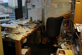 A cluttered office with a desk covered in papers, CDs, and miscellaneous items. A black office chair is positioned in front of the desk. There is a whiteboard on the wall with various notes scribbled in different colors. Shelves and boxes filled with more papers and folders surround the desk. The room has a window showcasing a cityscape with buildings in the background.