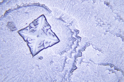 A close-up of a crystalline structure with intricate and sharp geometric patterns. The central feature is a diamond-shaped formation with jagged edges and a fine texture, suggesting a magnified view. Various shades of blue and white highlight the details, creating a visually striking contrast.