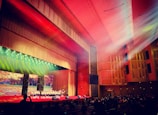 A large auditorium with wooden paneling and red carpeting is filled with an audience watching a group of performers on stage. The stage is brightly lit with colorful spotlights. The ceiling features intricate designs, and beams of light create a dramatic effect. A large screen behind the performers displays a nature scene with autumn leaves.