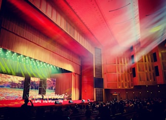 A large auditorium with wooden paneling and red carpeting is filled with an audience watching a group of performers on stage. The stage is brightly lit with colorful spotlights. The ceiling features intricate designs, and beams of light create a dramatic effect. A large screen behind the performers displays a nature scene with autumn leaves.