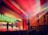 A large auditorium with wooden paneling and red carpeting is filled with an audience watching a group of performers on stage. The stage is brightly lit with colorful spotlights. The ceiling features intricate designs, and beams of light create a dramatic effect. A large screen behind the performers displays a nature scene with autumn leaves.