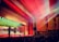 A large auditorium with wooden paneling and red carpeting is filled with an audience watching a group of performers on stage. The stage is brightly lit with colorful spotlights. The ceiling features intricate designs, and beams of light create a dramatic effect. A large screen behind the performers displays a nature scene with autumn leaves.