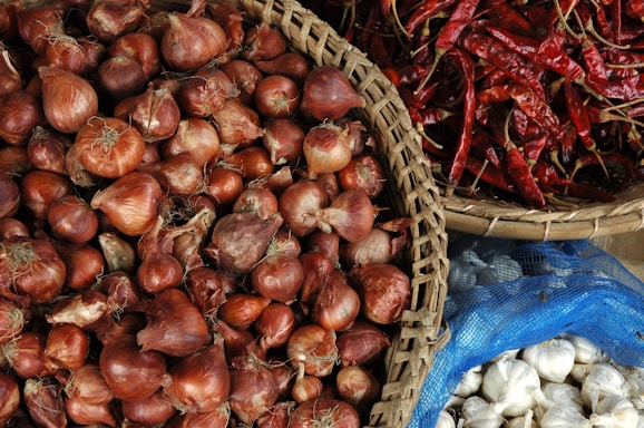 Baskets filled with fresh produce including red onions, dried red chilies, and white garlic cloves. The arrangement creates a vibrant and contrasting visual display.