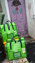 Two green Amazon Fresh delivery bags are placed on the ground in front of a uniquely painted purple door with decorative elements such as a crown and wings. The bags display various grocery items listed on them in white text. The surrounding area includes orange crates and a black mat, with the bags leaning against a wall.