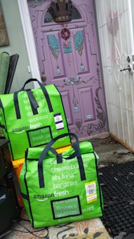 Two green Amazon Fresh delivery bags are placed on the ground in front of a uniquely painted purple door with decorative elements such as a crown and wings. The bags display various grocery items listed on them in white text. The surrounding area includes orange crates and a black mat, with the bags leaning against a wall.